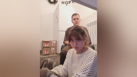 Media: Video of a young woman with brown hair in a white knit sweater sitting in a wheelchair, looking concerned. Behind her, a man in a brown shirt stands by a white refrigerator.