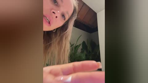 Media: A video of a blonde woman with fair skin, wearing hoop earrings, in a bathroom, looking slightly to the side. A blurred, partially visible male hand is in the foreground.
