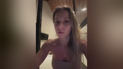 Media: A video of a blonde woman with a slender physique and fair skin, wearing a strapless beige dress, standing in a dimly lit room with wooden ceiling and blurred background.