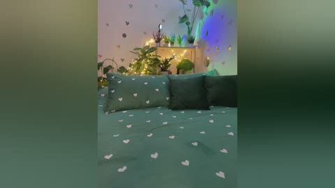 Video of a cozy, dimly-lit bedroom with a teal heart-patterned bedspread, green pillows, fairy lights, potted plants, and a wooden shelf adorned with small decor. The background features a soft, pastel-colored wall with scattered heart decals.