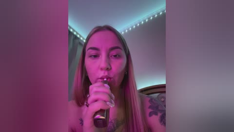 Media: Video of a young woman with long blonde hair, holding a glass pipe, in a dimly lit room with purple and blue lighting, showcasing tattoos on her arm.