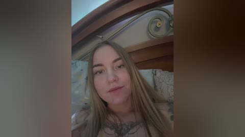 Media: Video of a young, fair-skinned woman with long, blonde hair, wearing a floral-patterned top, lying on a bed with a wooden headboard and a decorative pillow.
