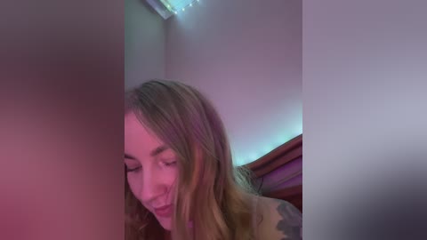 Media: Video of a smiling woman with shoulder-length light brown hair, wearing a brown jacket, in a dimly lit room with soft, multicolored lighting.