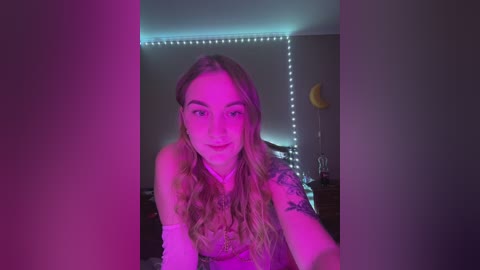 Media: A video of a young woman with long, wavy blonde hair and a tattoo on her left arm, smiling in a dimly lit bedroom with pink and purple lighting, string lights, and a yellow balloon.