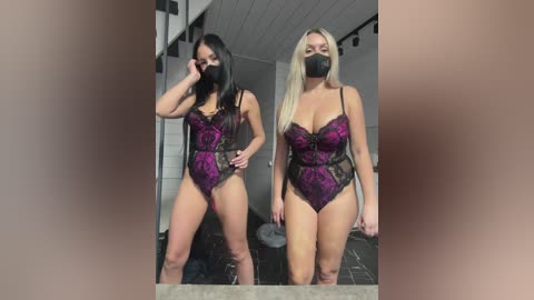 Media: Video of two women with fair skin, wearing matching purple and black lace bodysuits with masks, standing in a bathroom with grey tiles.