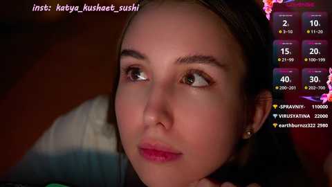 Media: Video of a young Asian woman with fair skin, straight brown hair, and light makeup, looking contemplative. Background includes a dimly lit room with a chat box overlay showing follower count and social media handles.