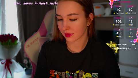 Media: A video of a young woman with fair skin and straight, brown hair, wearing a black graphic tee, and pink lipstick, indoors with a blurred background.