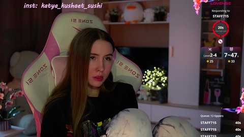 Media: Video of a young woman with long brown hair in a gaming chair, playing a video game, surrounded by plushies and a dimly lit room.