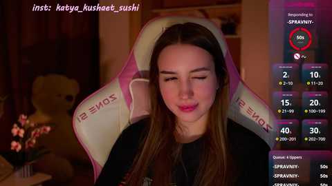 Media: Video of a young woman with long brown hair, wearing a black top, seated in a pink gaming chair. She has a neutral expression. Background includes a teddy bear and a dimly lit room.