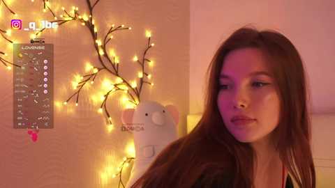 Video of a young woman with long brown hair, wearing a black jacket, standing beside a glowing string of yellow lights and a white elephant figurine on a beige wall.