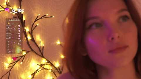 Video of a young woman with fair skin and reddish-brown hair, smiling softly, against a warmly lit, textured wall with a glowing string of fairy lights.