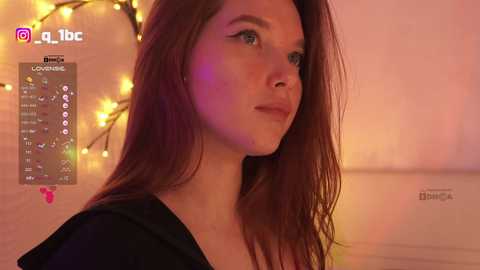 Video of a young Caucasian woman with fair skin and long brown hair, wearing a black top. Background features a cozy, warmly lit room with string lights and a brick wall. Social media icons are visible.