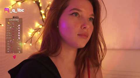 Video of a young woman with long brown hair, fair skin, and green eyes, wearing a black top, looking contemplative. Background features warm, ambient lighting and a string of fairy lights.