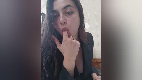 Media: A video of a young woman with long, dark hair, light skin, and full lips, provocatively licking her finger and seductively staring into the camera. She wears a black blazer and is seated against a wooden wall.