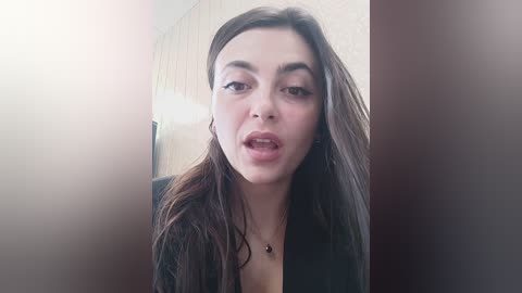 Media: A video of a young woman with long, dark hair, wearing a black top, and a necklace, making a funny face, indoors with a blurred background.