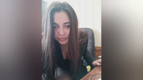 Media: Video of a young woman with long dark hair and fair skin, wearing a black top, sitting at a desk in a dimly lit office.