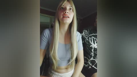 Video of a young woman with long blonde hair, wearing a light blue crop top and white lace skirt, standing indoors against a dark background.