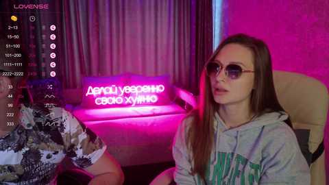 A video of a young woman with long brown hair, wearing sunglasses and a gray hoodie, seated next to a man in a camouflage shirt, in a dimly lit room with neon signs and pink lighting.