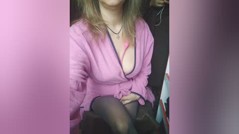 Media: Video of a light-skinned woman with medium-length brown hair, wearing a pink robe with a black lace bra visible, sitting on a dark chair.