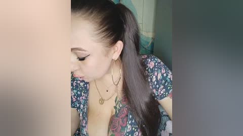 A video of a young Asian woman with long, dark hair in a high ponytail, wearing a floral-patterned blouse, hoop earrings, and a necklace, sitting in a dimly lit room.
