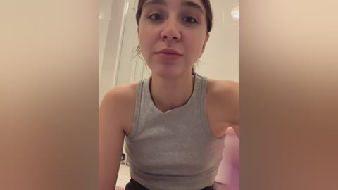 Media: Video of a young woman with fair skin, brown hair in a ponytail, wearing a grey tank top, standing in a bathroom with beige walls and a pink bottle.