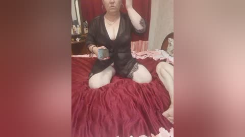 Video of a middle-aged woman with light skin, wearing a sheer black negligee, sitting on a red bedspread, holding a cup, in a dimly lit bedroom.