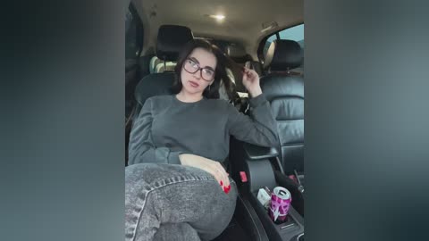 Media: Video of a young woman with fair skin, glasses, and dark hair, wearing a gray sweatshirt and jeans, reclining in a car seat, holding a purple drink can.