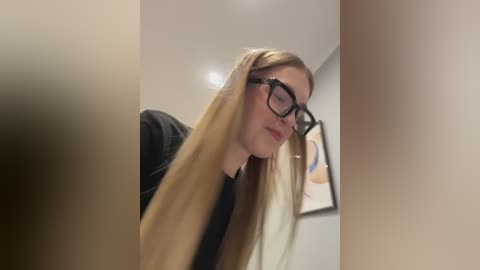 Media: Video of a fair-skinned woman with long, straight blonde hair, wearing black-framed glasses and a black top, standing indoors with beige walls and a framed artwork in the background.