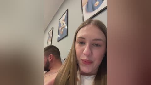 Media: Video of a young Caucasian woman with long blonde hair, wearing a white top, standing in a hallway with framed abstract art on light grey walls, partially obscured by a man in the background.