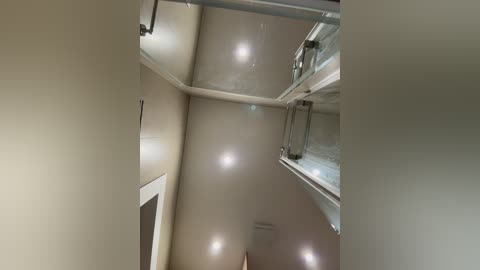 Video of a modern interior space with beige walls and a high ceiling. The ceiling features recessed lights and a glass staircase railing on the right. The left wall has a white-framed door. The scene is clean and minimalist.