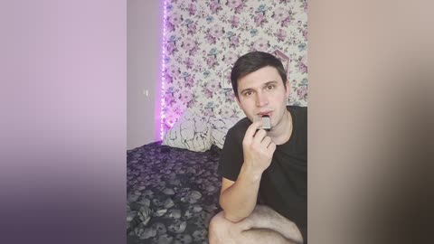 A video of a young man with short dark hair, wearing a black t-shirt, sitting on a bed with a dark floral-patterned blanket. He is holding a cigarette to his lips. The background features a floral tapestry with purple lights.