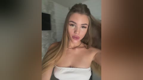Video of a young, light-skinned woman with long, straight, blonde hair. She has a slender build and is wearing a strapless white top. The background features a white brick wall and a flat-screen TV.