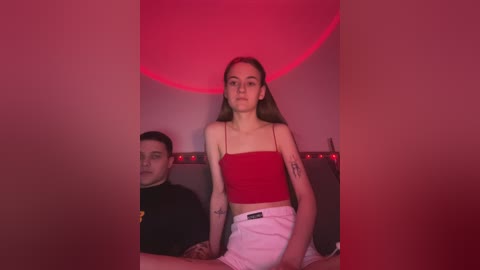 Media: Video of a young woman with long brown hair in a red crop top, white shorts, and tattoos, sitting next to a man in a black hoodie, in a dimly lit room with red lighting.