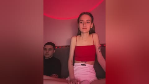 Media: Video of a young woman with fair skin and straight brown hair, wearing a red crop top and white shorts, standing against a red-lit wall. A man with dark hair and a black turtleneck sits behind her.