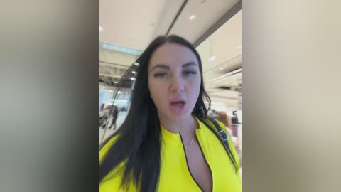 Video of a young woman with long black hair, fair skin, wearing a bright yellow jacket, standing in a modern indoor space with glass walls and ambient lighting.
