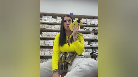 Video of a woman with long black hair, wearing a bright yellow shirt and gray pants, taking a selfie in a pharmacy with numerous medicine boxes in the background.