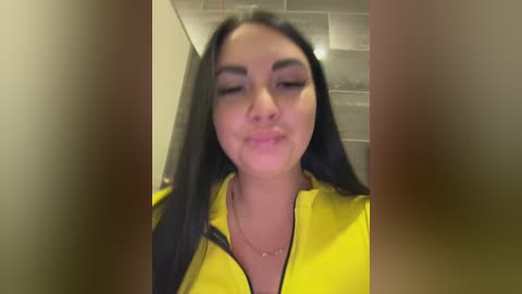 A video of a young woman with long black hair and fair skin, wearing a yellow jacket, blowing a kiss in a bathroom with gray tiled walls.