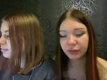 Video of two young women with long hair, one with straight brown hair, the other with straight brown hair with red streaks, both wearing black leather jackets. Dark wallpaper with intricate patterns in the background.