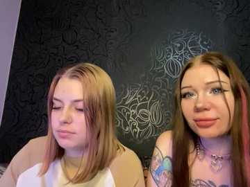 Video of two young women with long, straight hair, one in a white top, the other in a pink top, both with tattoos, in front of a dark, patterned wall.