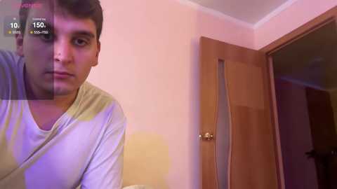 Media: A video of a young man with short dark hair, wearing a white t-shirt, standing in a dimly lit room with a closed wooden door and pale pink walls.