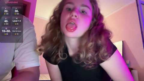 Media: Video of a young woman with curly hair, sticking out her tongue, wearing a black top, in a dimly lit bedroom.