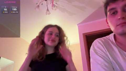Media: Video of a young woman with curly hair and a young man with short hair, both in casual attire, indoors under pinkish lighting.