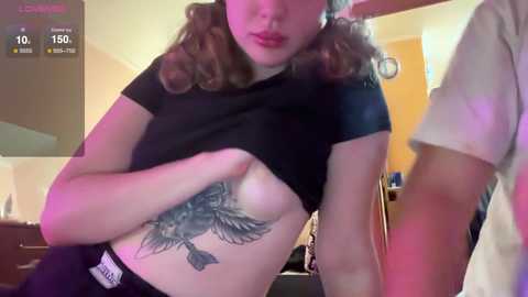 Media: Video of a woman with wavy brown hair, lifting her black shirt to reveal a detailed black-and-gray tattoo of a bird on her left side. She stands in a dimly lit room with a purple hue.
