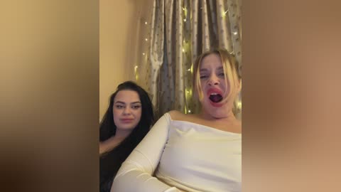 Video of two women, one with long black hair and the other with blonde hair, both wearing off-shoulder white tops, smiling and sticking out their tongues, against a background of beige curtains with fairy lights.