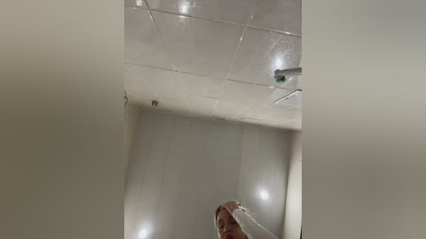 Video of a woman in a bathroom with white tiled walls and ceiling, holding a towel, wearing a pink robe, and standing in front of a mirror.