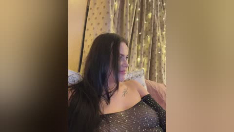 A video of a woman with long, dark hair, wearing a black sequined off-shoulder dress, sitting on a bed with patterned curtains in the background.