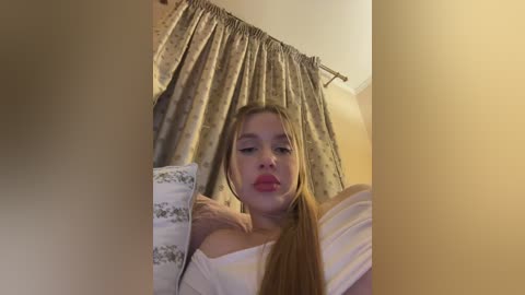 Video of a young woman with fair skin, light brown hair, and full lips, wearing a white off-shoulder top, standing in a bedroom with beige walls and floral-patterned curtains.