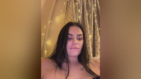 Video of a topless, fair-skinned woman with long dark hair and a slender physique, wearing dark lipstick. She stands in front of a golden curtain with fairy lights, smiling subtly.