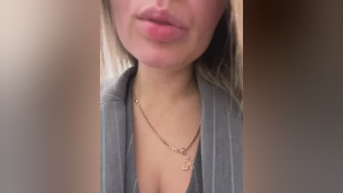 Media: Video of a woman with light skin, wearing a grey cardigan over a low-cut top, revealing cleavage. She has straight, shoulder-length blonde hair, a gold necklace with a heart pendant, and full lips. The background is blurred, with neutral tones.
