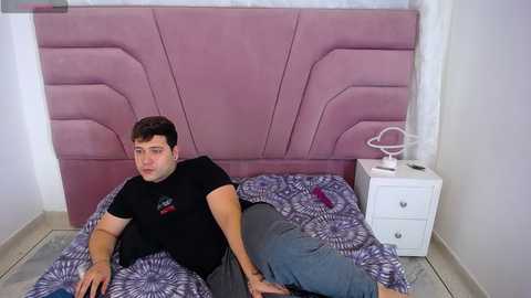 Video of a man with short dark hair, wearing a black t-shirt and gray sweatpants, lying on a bed with a lavender upholstered headboard and purple and white patterned sheets. A white nightstand with a round lamp and a book is visible to the right.